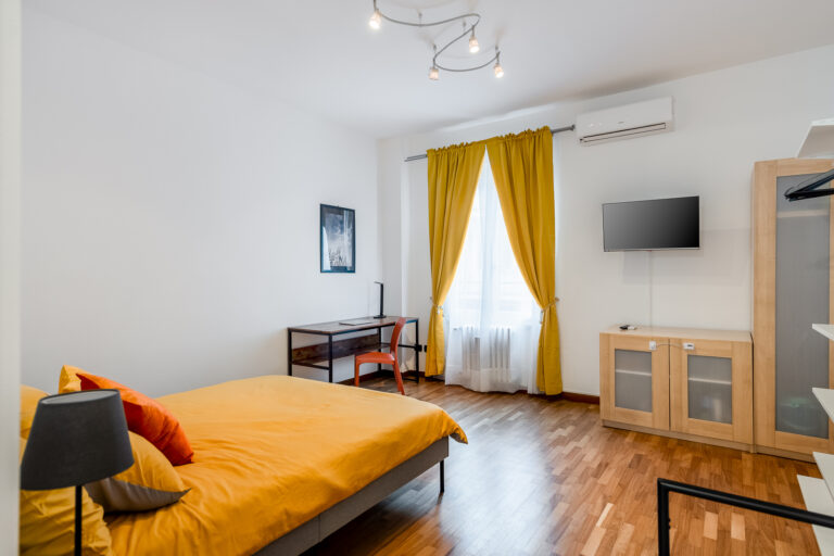 Habitación moderna con cama de sábanas amarillas, escritorio con silla naranja, cortinas doradas, TV y aire acondicionado.