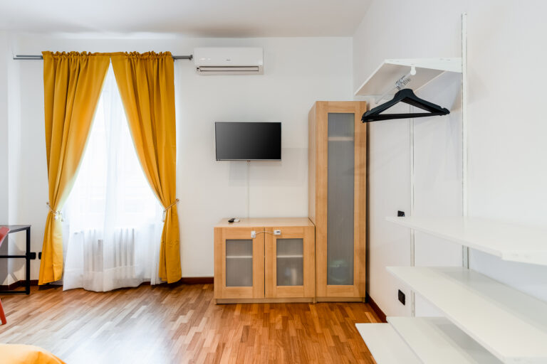 Habitación luminosa con cortinas amarillas, TV montada en la pared, armario de madera y estanterías blancas. Piso de madera clara.