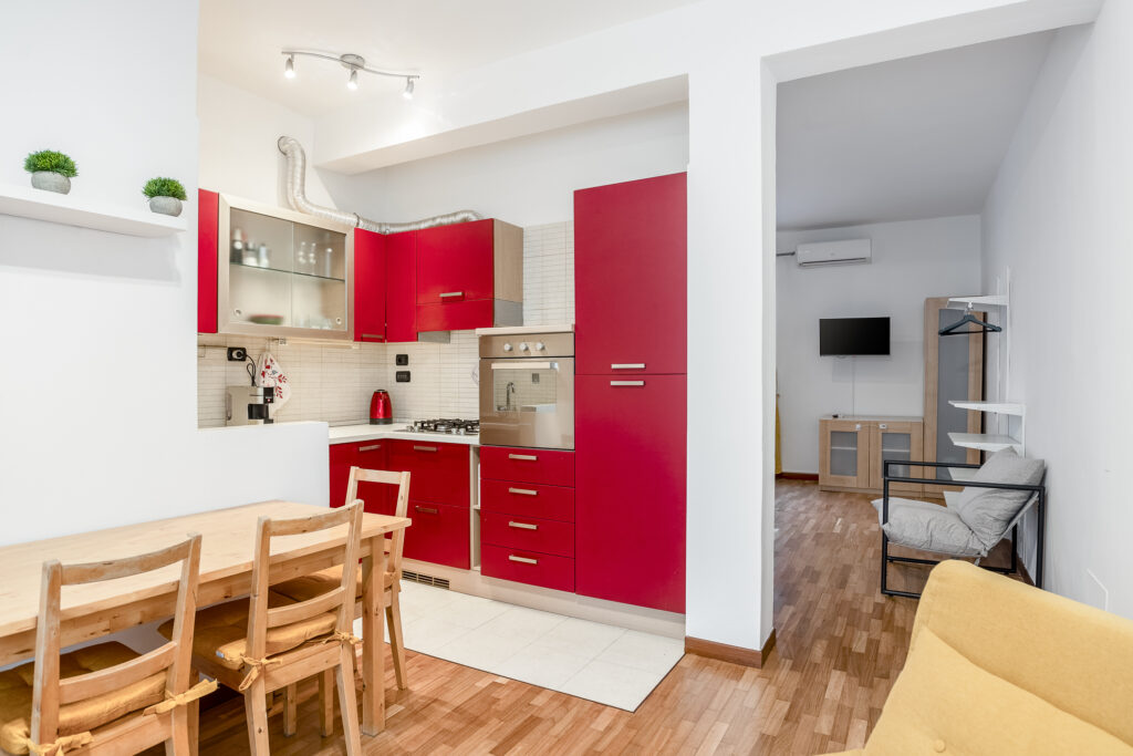 Cocina moderna con gabinetes rojos, mesa de madera con sillas, y sofá gris. Área de sala con televisor y aire acondicionado.
