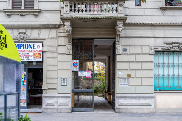 Edificio antiguo de piedra con puerta de entrada de hierro forjado, balcón decorativo y pizzería a la izquierda.