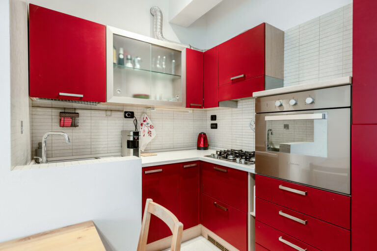 Cocina moderna con gabinetes rojos, horno empotrado, vitrocerámica, fregadero y electrodomésticos sobre encimera blanca.
