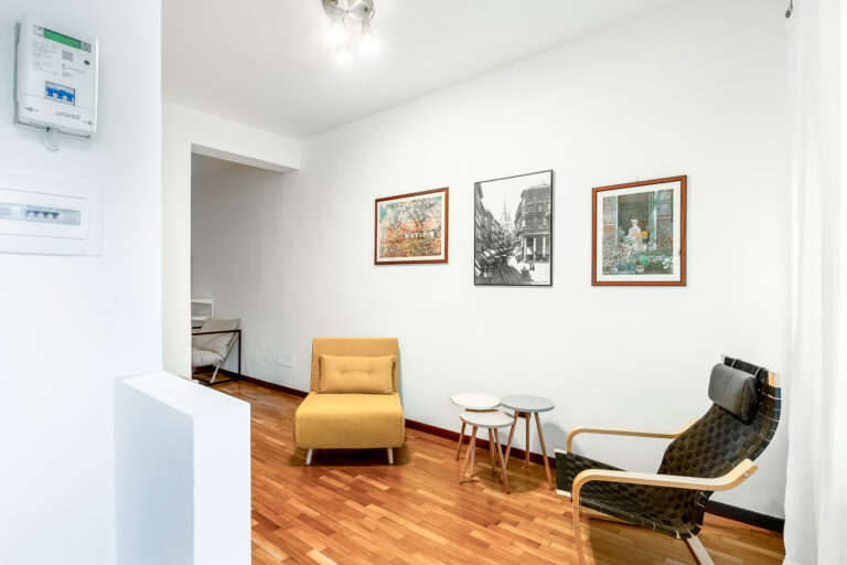 Sala con pared blanca, tres cuadros, sillón amarillo, silla negra, mesa con tres mesas pequeñas, suelo de madera y luz de techo.