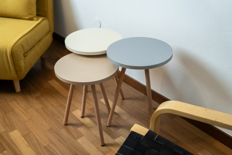 Tres mesas redondas apiladas en tonos gris, beige y crema, con patas de madera clara, junto a un sofá amarillo y una silla de madera.