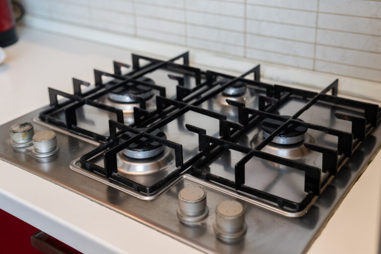 Estufa de gas de cuatro quemadores con parrillas negras, perillas metálicas y fondo de azulejos blancos.