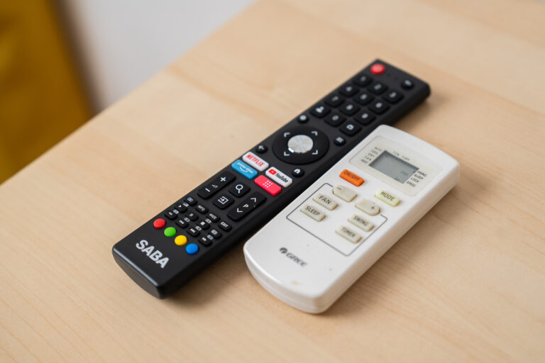 Dos controles remotos sobre mesa de madera: uno negro con botones coloridos y etiquetas de streaming, otro blanco con pantalla LCD.