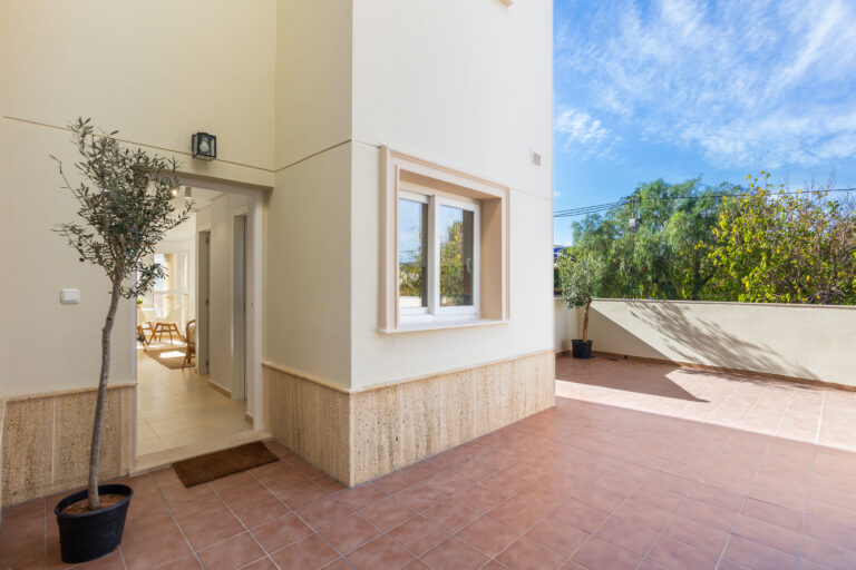 Moderna entrada a casa con piso de baldosas rosadas, planta en maceta, ventana grande y árboles al fondo bajo cielo azul.