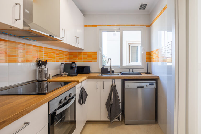 Cocina moderna con encimeras de madera, azulejos naranjas, horno, fogón, fregadero y lavavajillas plateado, luz natural.