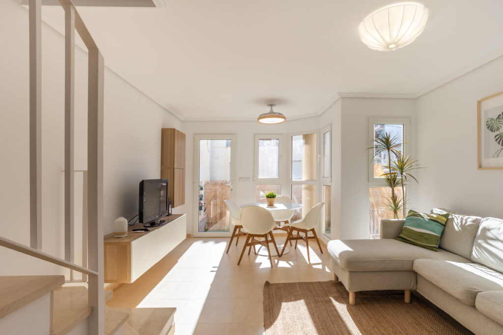 Sala de estar luminosa con sofá gris, mesa redonda blanca y sillas. Plantas y decoración moderna. Ventanales amplios y suelo claro.