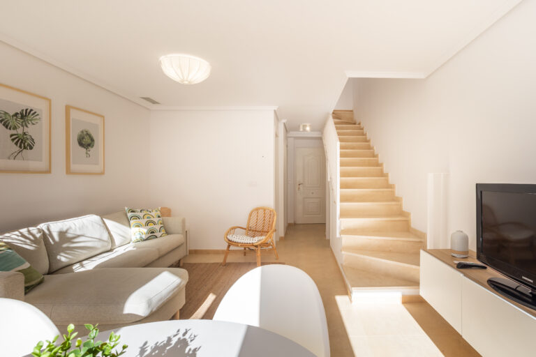 Sala luminosa con sofá beige, cojines estampados, silla de mimbre, escalera beige y TV en mueble blanco. Iluminación suave.