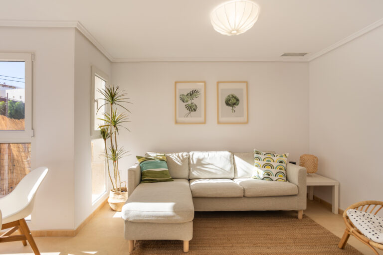 Sala de estar con sofá beige, cojines verdes y estampados, planta alta en esquina, cuadros de hojas, luz natural.