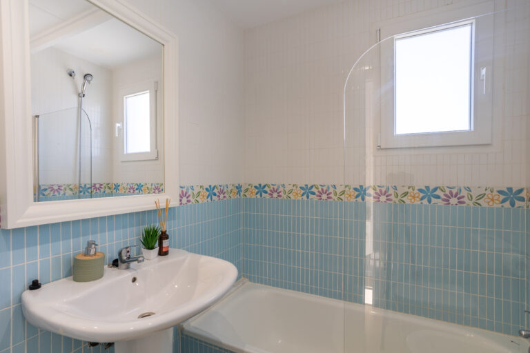 Baño con azulejos azul claro y borde de flores coloridas. Espejo grande, lavabo blanco y ducha al fondo. Ventana pequeña y decoración sencilla.