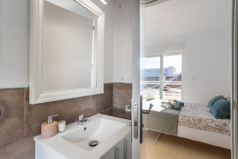 Baño moderno con lavabo blanco, espejo grande, dispensador de jabón y vela. Vista a un dormitorio con cama decorada y ventana soleada.