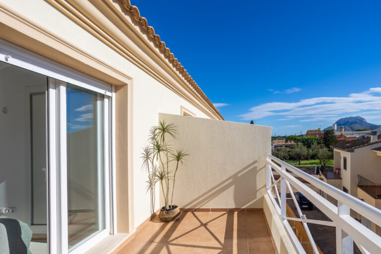 Balcón con piso de cerámica, planta en maceta, barandal blanco; cielo azul, casas y montaña al fondo.
