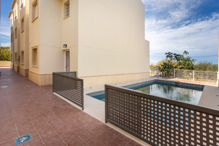 Edificio beige con piscina rectangular cercada, pavimento de baldosas marrones, cielo azul y vegetación al fondo.