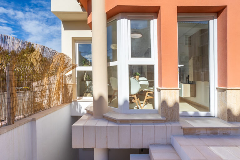 Ventana grande en pared de ladrillo naranja, interior con mesa blanca y sillas. Jardín exterior con cañas y cielo azul claro.