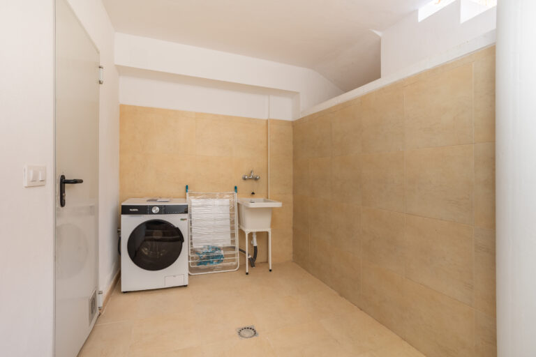 Lavandería con lavadora moderna, fregadero blanco, azulejos beige en paredes y suelo, y tendedero plegable.