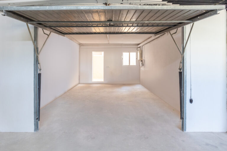 Garaje vacío con piso de concreto, paredes blancas y puerta abierta. Ventana y puerta al fondo, techo metálico.