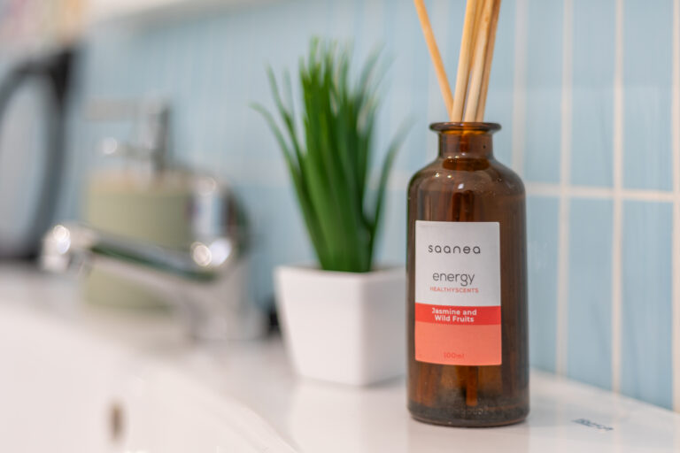 Botella de fragancia, etiqueta "saanea energy, Jasmine and Wild Fruits", al lado de un lavabo y una planta en maceta.