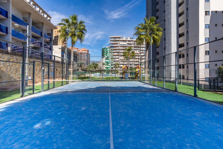 Pista de pádel azul rodeada de edificaciones altas. Palmeras y zona de juegos al fondo bajo un cielo despejado.