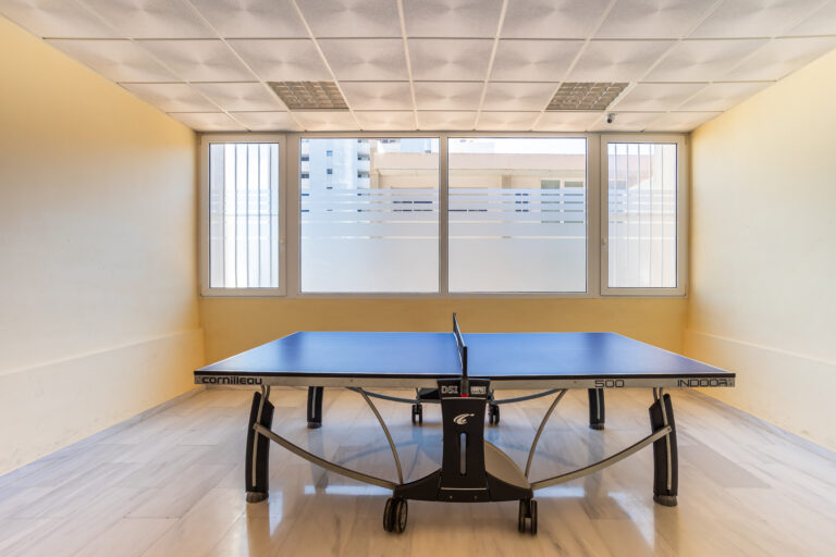 Mesa de ping-pong azul en una sala vacía con paredes amarillas, ventanas grandes y suelo de mármol claro.