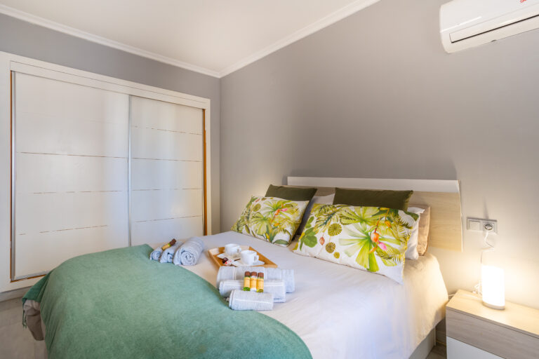 Dormitorio con pared gris, cama doble con cojines tropicales, mantas verdes, armario blanco y lámpara encendida en el buró.