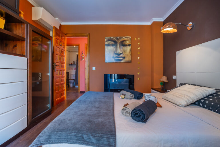 Habitación con paredes marrones, cama con sábanas grises, toallas enrolladas, arte de rostro dorado y puerta hacia el baño.