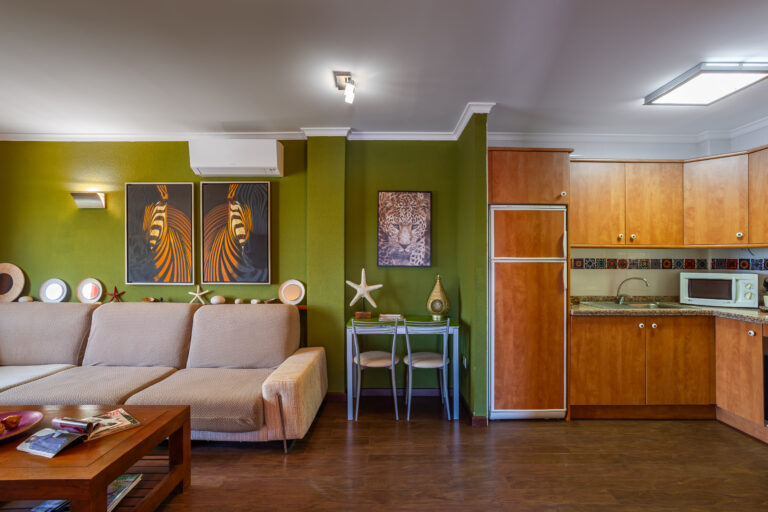 Sala con sofá beige, pared verde, arte de animales, mesa pequeña, cocina de madera clara, electrodomésticos modernos.