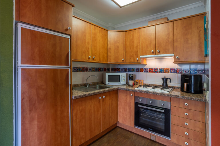 Cocina con gabinetes de madera clara, encimera de granito, microondas, cafetera, estufa con horno y azulejos decorativos.