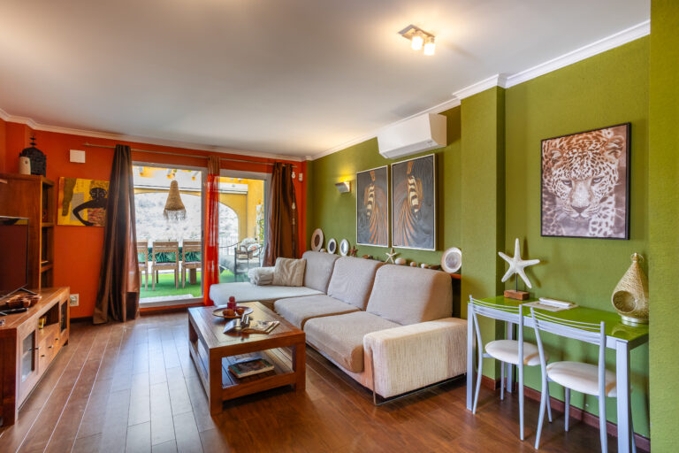 Sala acogedora con paredes verdes y naranjas, sofás beige, mesa de madera, arte de animales y gran ventanal hacia un patio.