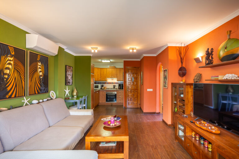 Sala de estar colorida con paredes verdes y naranjas, sofá beige, mesa de madera, arte en las paredes y cocina al fondo.