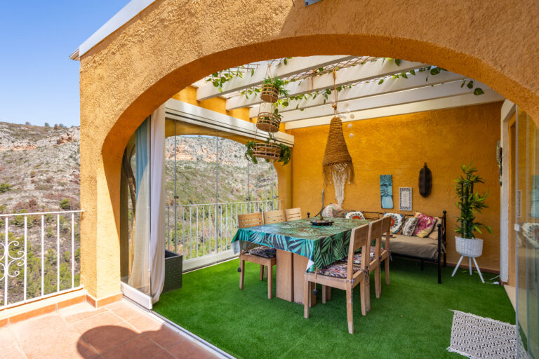 Terraza cubierta con arcos, paredes amarillas, mesa decorada, sillas de madera, plantas colgantes y suelo de césped artificial. Vista a montañas.