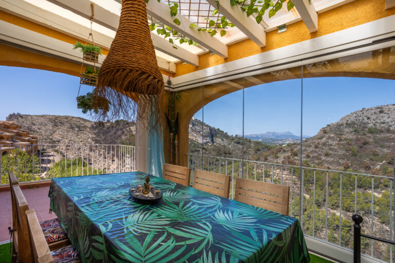 Comedor al aire libre con mesa grande cubierta con mantel de hojas verdes. Vista panorámica a montañas y cielo despejado.