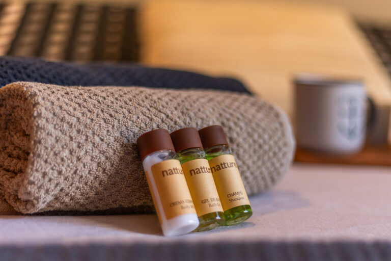 Toallas enrolladas y botellas pequeñas de champú, gel y crema sobre una cama. Taza difusa de fondo.