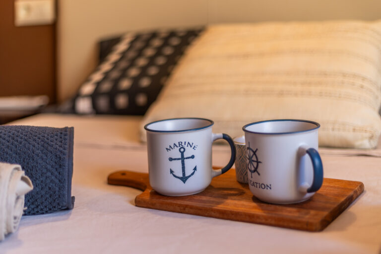 Tazas blancas con temática marina sobre tabla de madera, fondo con cojines y toalla enrollada.
