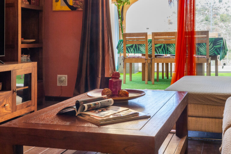 Sala de estar con mesa de madera, revistas, cojín beige y cortinas. Mesa de comedor en terraza con mantel verde.