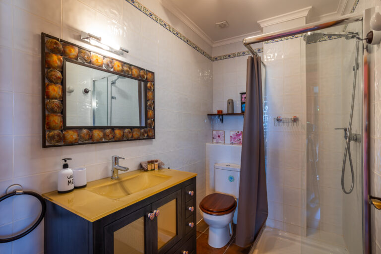 Baño luminoso con lavabo amarillo, espejo decorativo, ducha con cortina gris y estante sobre el inodoro.