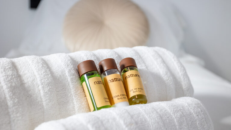 Botellas de cortesía con champú, crema corporal y gel de baño sobre toallas blancas en una habitación.