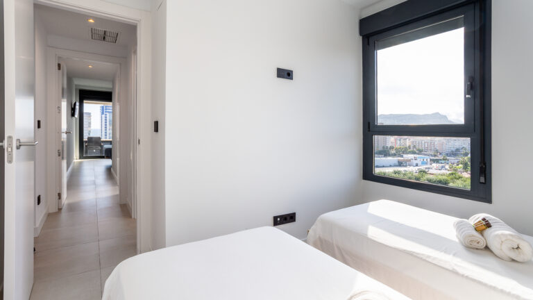 Pasillo con suelo de baldosas conecta dos habitaciones. Ventana grande con vista a edificios y montañas. Camas blancas y toallas.