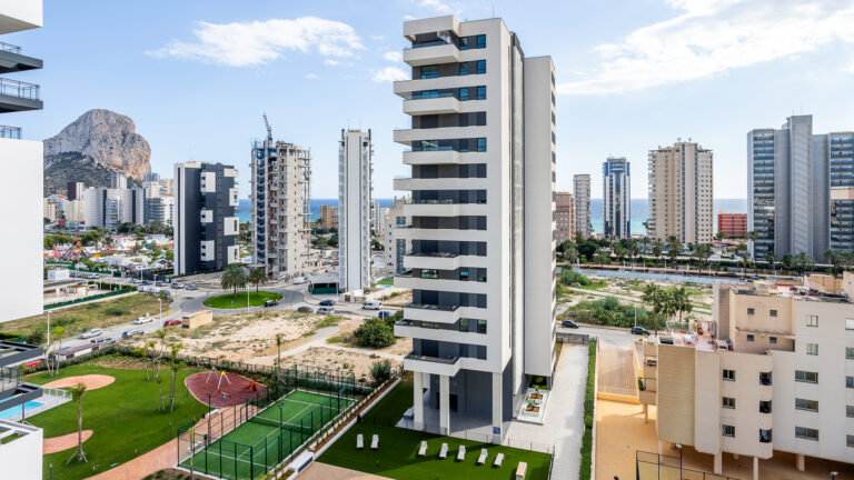 Edificios modernos con balcones, parque infantil abajo, mar de fondo y una montaña a la izquierda bajo cielo azul.