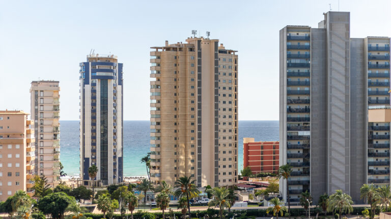 Edificios altos frente al mar con balcones, cielo despejado y palmeras en primer plano.