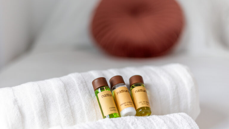 Botellas de champú, crema corporal y gel de baño sobre toalla blanca. Fondo difuminado con cojín marrón.