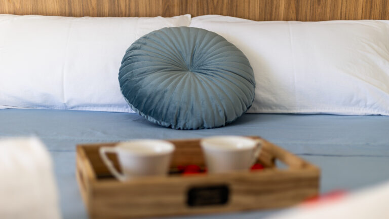Almohada azul redonda sobre cama con sábanas azules y almohadas blancas. Bandeja de madera con dos tazas al frente.