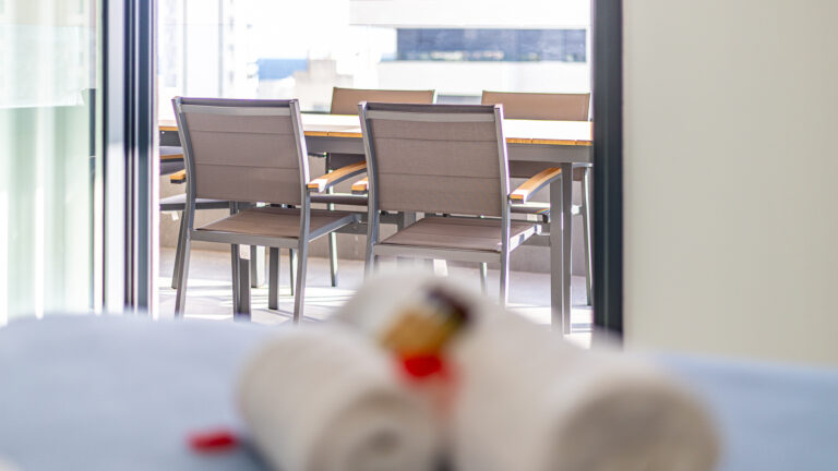 Sillas de metal y tela beige alrededor de una mesa en un balcón luminoso. Toallas enrolladas en primer plano.