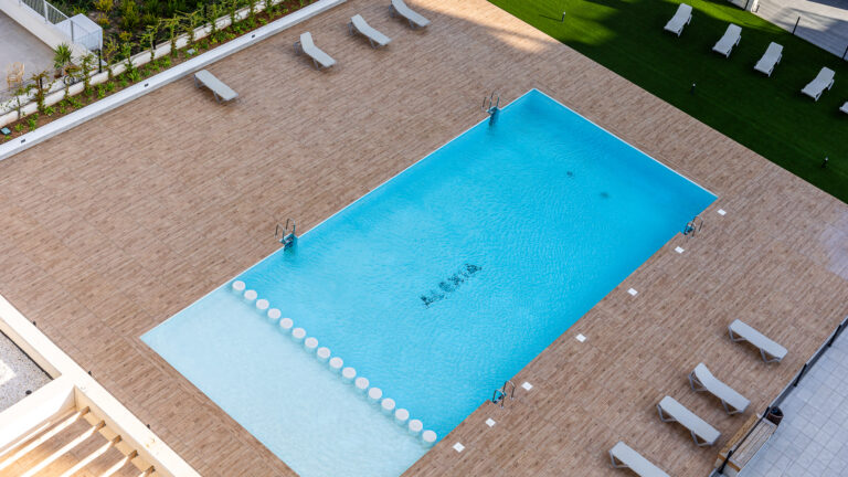 Piscina rectangular azul con borde de madera; tumbonas grises alineadas; césped al fondo; ambiente despejado.