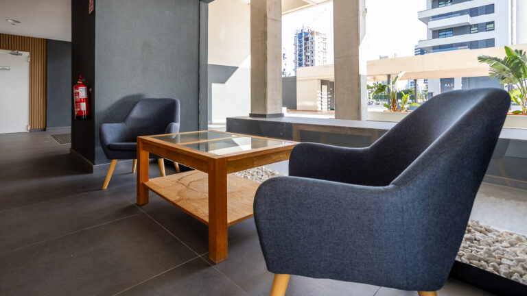 Zona moderna de descanso con dos sillones grises y mesa de madera. Edificios de fondo, plantas y decoraciones en gris.