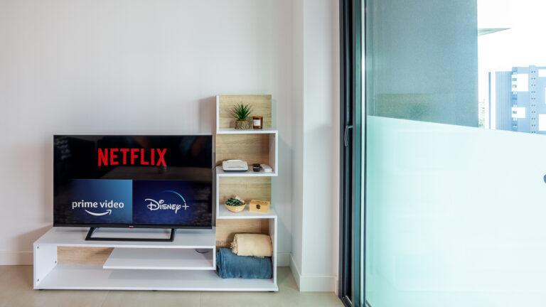 Televisor con logos de Netflix, Prime Video y Disney+ en mueble blanco. Planta, router y mantas al lado. Ventana de vidrio a la derecha.