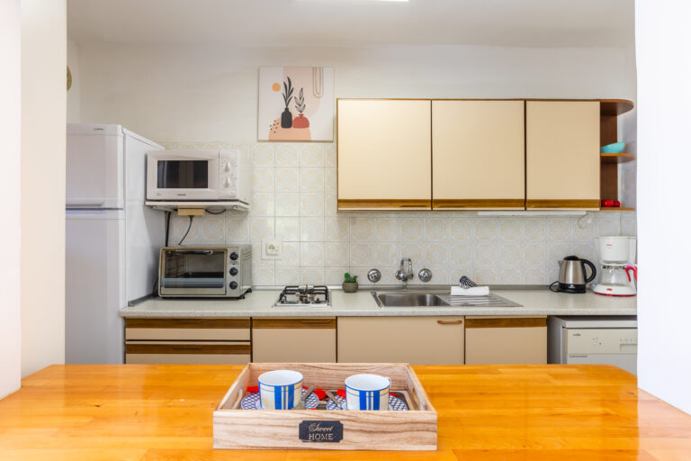 Cocina con muebles beige, microondas, horno, fregadero y cafetera. Azulejos decorativos en la pared y bandeja con tazas.