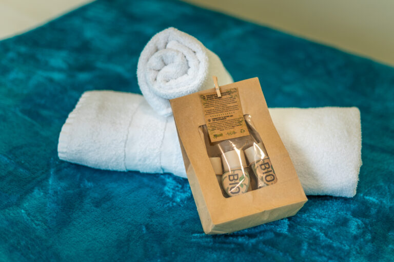Toallas blancas enrolladas sobre manta azul y kit de baño bio en empaque marrón transparente.