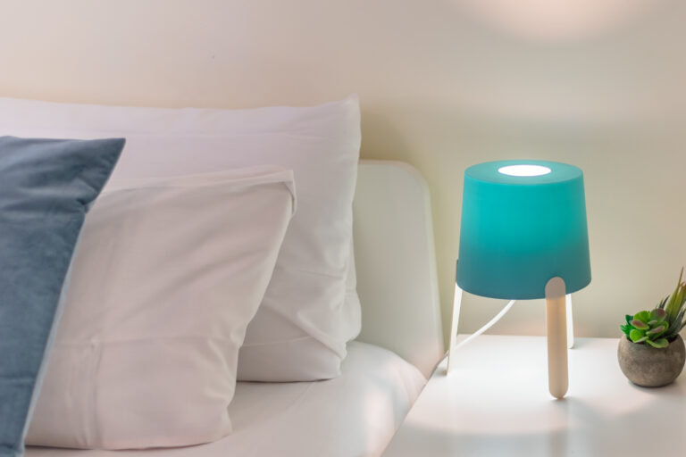 Lámpara azul brillante sobre mesita blanca junto a cama con sábanas blancas y cojín azul, planta en maceta gris.