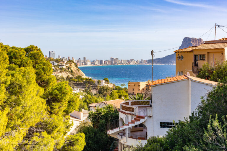 Costas rocosas con casas blancas y tejados naranjas, mar azul y una ciudad al fondo bajo un cielo despejado.